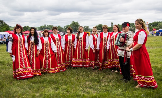 A group of people dressed in traditional red folk costumes stand in a semicircle outdoors on a grassy field. The women wear long red dresses with intricate gold and white patterns, while a man holds an accordion. The scene suggests a cultural or traditional gathering, possibly a festival or celebration. Tents and trees are visible in the background under a cloudy sky.