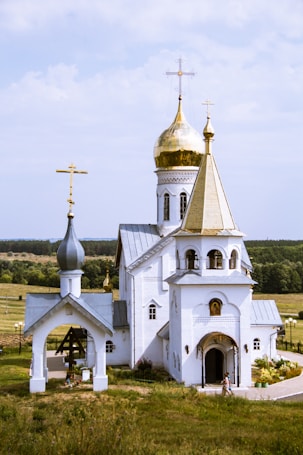 A picturesque Orthodox church with white walls and golden domes sits in a pastoral landscape. The architecture features arches and crosses, and a single person walks near the entrance. Surrounding the church is a well-kept garden and path, with fields and forests in the background, all under a partly cloudy sky.