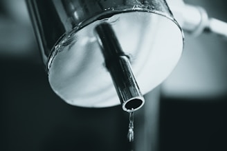 A close-up view of a metal faucet with water dripping from the spout. The metal surface has a reflective quality, and the image is captured in a monochromatic color palette.