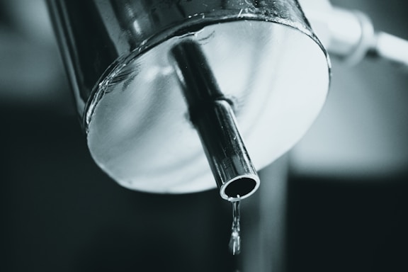 A close-up view of a metal faucet with water dripping from the spout. The metal surface has a reflective quality, and the image is captured in a monochromatic color palette.