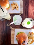 A wooden table holds two wooden serving boards, each with a burger and a side of potato wedges. Three different beverages in tall glasses are present, garnished with lime and mint, and varying in straw colors.