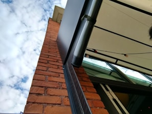 A brick wall extends vertically against a backdrop of a partly cloudy blue sky. A modern, metallic awning with a white canopy and some outdoor string lights is attached to the wall. The scene is viewed from an upward angle, giving a sense of height and openness.