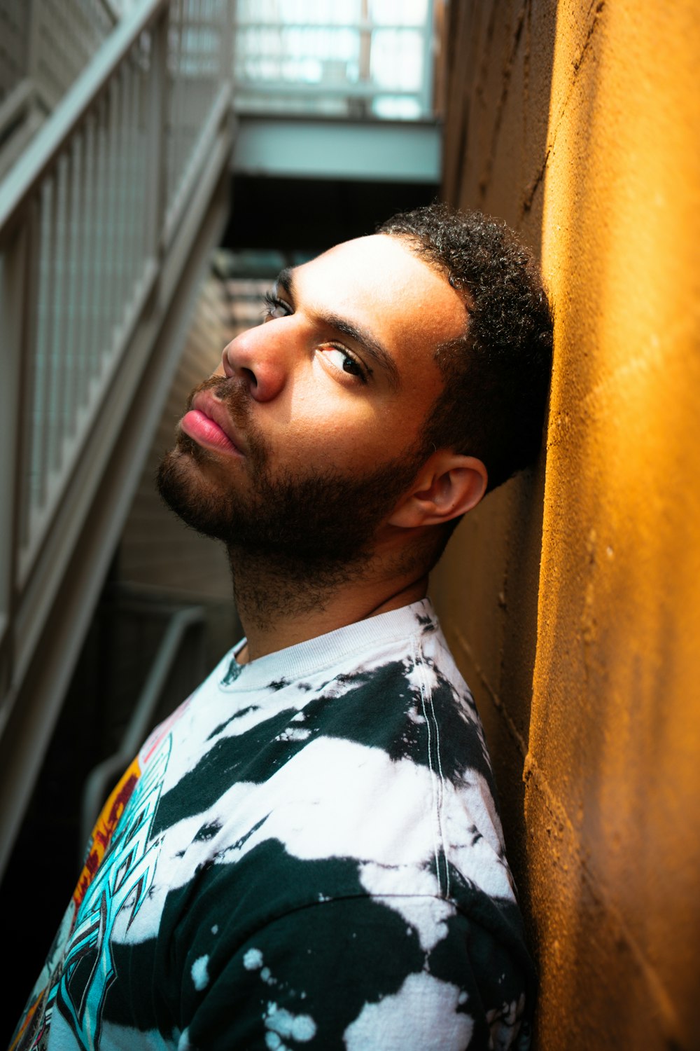 man in white black and green floral shirt leaning on yellow wall