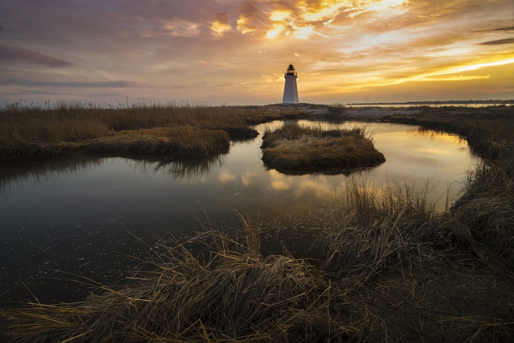 Weißer Leuchtturm auf braunem Grasfeld in der Nähe von Gewässern während des Sonnenuntergangs