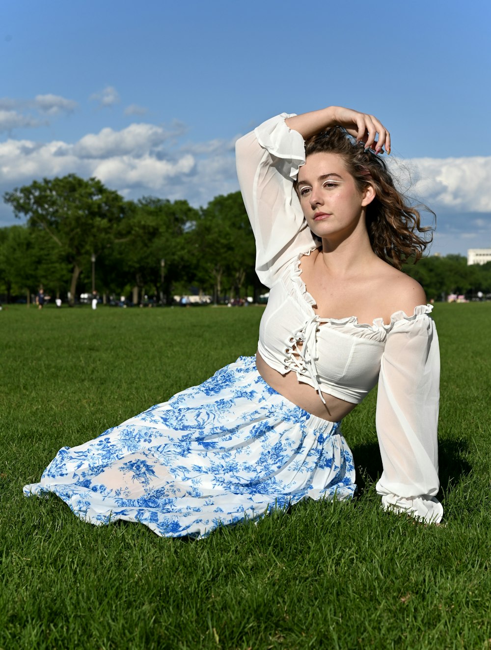 青空の下の緑の草原に横たわっている白と青の花柄のドレスを着た女性