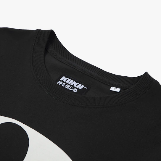 A close-up of a t-shirt featuring a minimalist badge logo in muted charcoal on a washed black fabric background.
