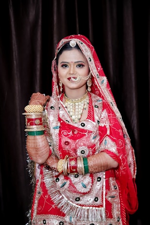 A woman dressed in traditional Indian bridal attire, adorned with intricate henna designs on her arms. She is wearing a vibrant red sari with elaborate embroidery and patterns, along with multiple golden and colorful bangles. The jewelry includes a nose ring, earrings, and a necklace, complementing her ornate outfit.