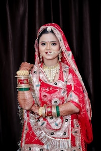 A woman dressed in traditional Indian bridal attire, adorned with intricate henna designs on her arms. She is wearing a vibrant red sari with elaborate embroidery and patterns, along with multiple golden and colorful bangles. The jewelry includes a nose ring, earrings, and a necklace, complementing her ornate outfit.