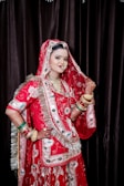 A woman dressed in traditional South Asian bridal attire, featuring an elaborately embroidered red garment with intricate designs. She is adorned with multiple pieces of jewelry, including necklaces, earrings, and bangles, and her hands are decorated with henna. The background is a dark curtain that contrasts with the vibrant colors of her outfit.