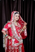 A woman dressed in traditional South Asian bridal attire, featuring an elaborately embroidered red garment with intricate designs. She is adorned with multiple pieces of jewelry, including necklaces, earrings, and bangles, and her hands are decorated with henna. The background is a dark curtain that contrasts with the vibrant colors of her outfit.
