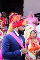 A man in a dark suit with a colorful turban and a pink scarf is standing among a group of people dressed in vibrant traditional attire. The women in the background are wearing sarees in various bright colors, indicating a festive or cultural event.