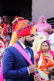 A man in a dark suit with a colorful turban and a pink scarf is standing among a group of people dressed in vibrant traditional attire. The women in the background are wearing sarees in various bright colors, indicating a festive or cultural event.