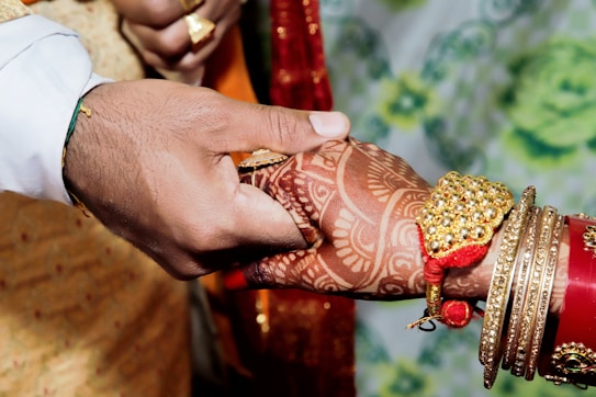 A close-up of two hands holding each other, adorned with intricate henna designs and jewelry. One hand wears gold bangles and a decorative wristband, while the other hand is holding it gently. The background is softly blurred with hints of green and blue.