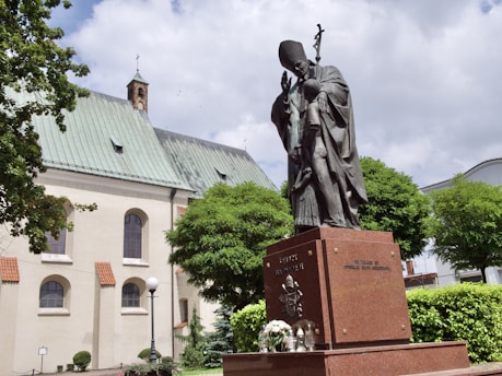 A large bronze statue of a religious figure stands on a granite pedestal in front of an old church building. The statue holds a cross and appears to be in a blessing pose. The surroundings include lush green trees, a lamp post, and a partly cloudy sky.