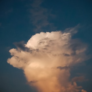 A large, towering cloud with a fluffy texture rises against a deep blue sky. The cloud is illuminated with warm, golden hues, creating a dramatic contrast with the surrounding sky.