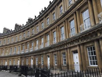 A row of connected Georgian-style townhouses with uniform architecture featuring tall windows and classic columns. The structure is curved, displaying intricate detailing above the windows and doors. Black wrought iron fences line the walkway in front.