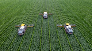 Three agricultural machines are evenly spaced across a vast, lush green field, likely a crop such as corn or maize. The machines are equipped with sprayers and appear to be engaged in agricultural work, perhaps spraying fertilizers or pesticides. The rows of crops are neatly aligned, creating a pattern that emphasizes the scale and organization of the farmland.