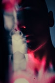 A double exposure portrait featuring the silhouette of a face with dramatic lighting. The interplay of shadows and pinkish-red hues creates a mysterious and abstract appearance. The image blends facial features with a soft, diffused background.