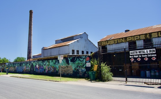 An industrial building with a rusted roof and a tall chimney stands adjacent to a colorful, graffiti-covered wall. The scene includes signs directing to Austin Metal & Iron. A clear blue sky serves as a backdrop to this urban setting.