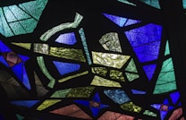 A vibrant stained glass artwork featuring geometric shapes and a variety of colors. The pieces are arranged in an abstract pattern, with overlapping layers of textured glass creating dynamic visual interest.