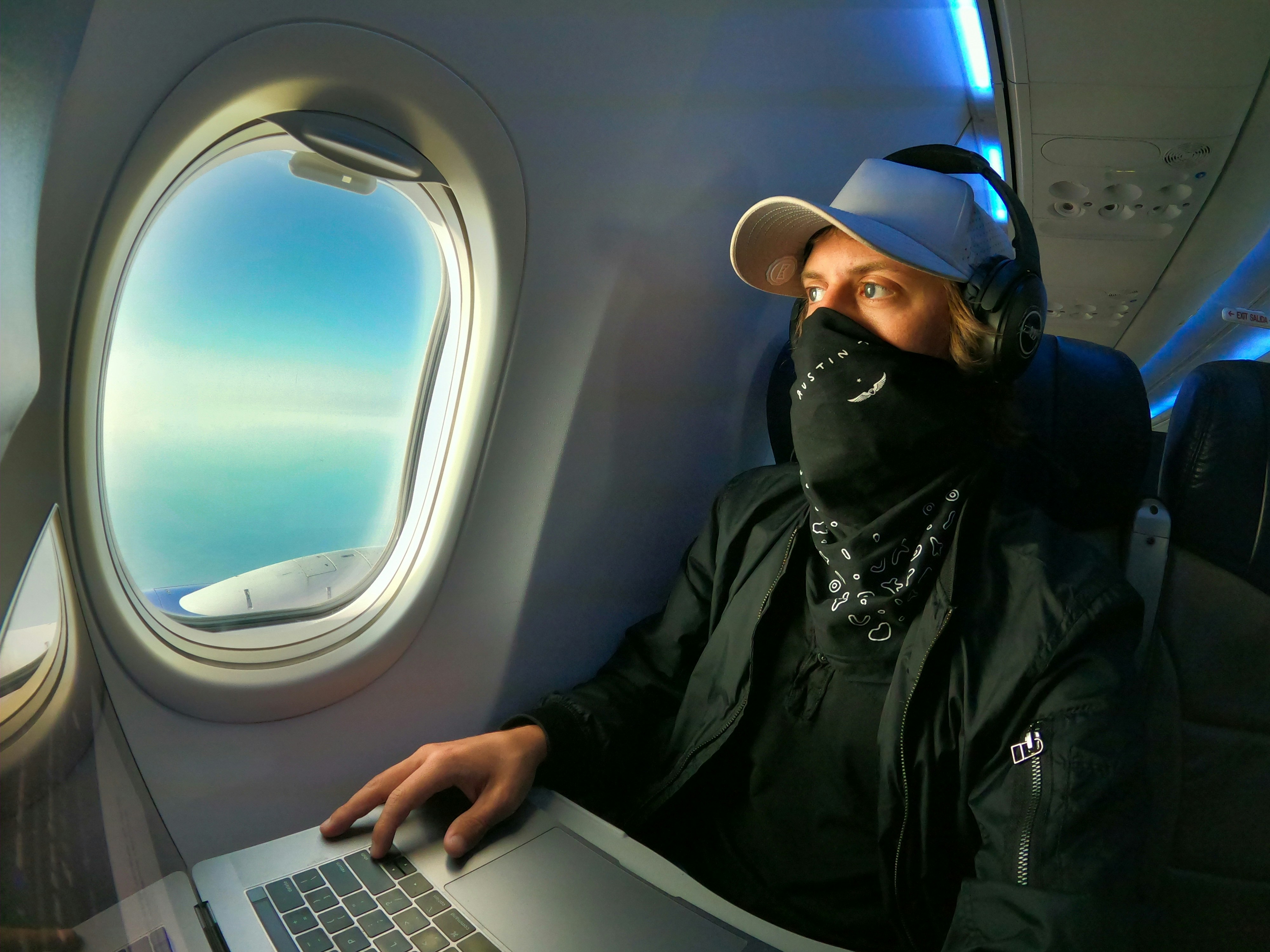 Digital nomad working remotely on his laptop while flying on an airplane looking out from his window seat at the Caribbean sea. Man is wearing a mask to travel safely for covid.