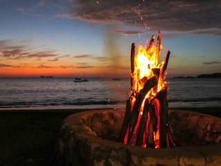 Evening bonfire on the beach with yachts visible in the background.