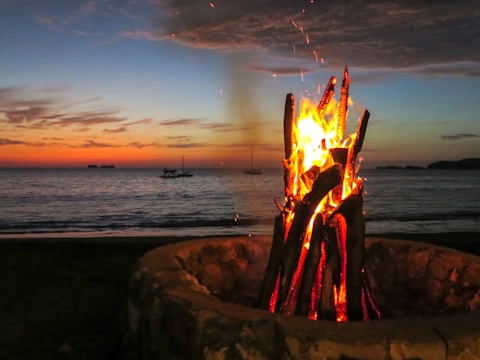Evening bonfire on the beach with yachts visible in the background.