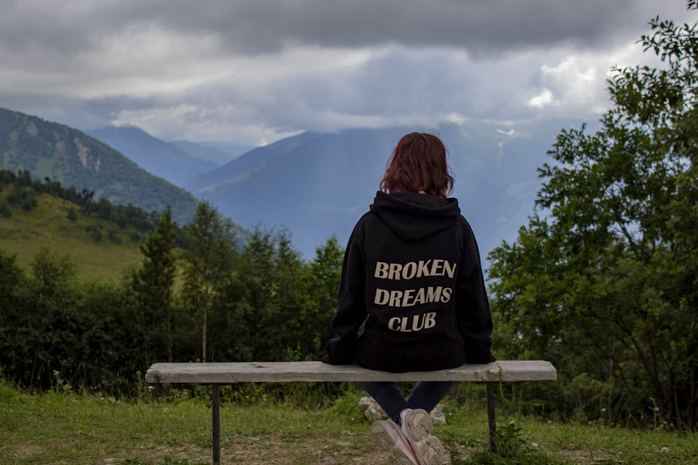woman in black and white hoodie sitting on brown wooden bench looking at mountains during daytime