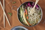 A bowl of noodles in broth is topped with thinly sliced vegetables such as zucchini, carrots, and red cabbage, garnished with sprouts. A pair of chopsticks rests on top of the bowl, poised to pick up the noodles. The scene is set on a brown fabric background, with a second pair of chopsticks placed beside the bowl, along with a few scattered greens.