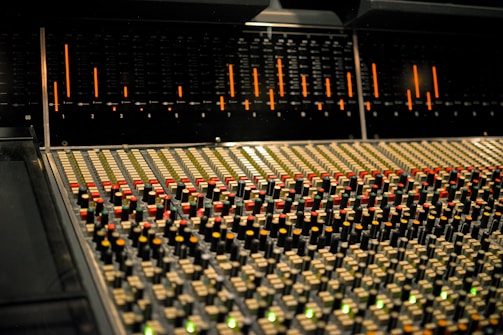 A professional mixing console with numerous buttons, faders, and knobs illuminated by soft lighting. The control panel features a complex array of colors, primarily orange and green, used for audio mixing in a studio environment.