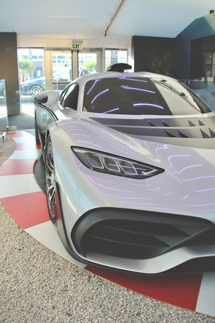 A sleek, futuristic sports car with a silver body is displayed indoors. The car has a low profile, sharp contours, and high-performance design features. The environment includes a modern showroom with glass walls, allowing natural light to filter in. The floor around the car has a red and white checkered pattern.