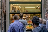Several people stand outside a window counter where pizzas are being sold. The vendor holds a piece of paper and is engaging with the customers. Various pizzas are displayed on the counter, and one has a sign that reads 'Venezia 15.00'. There are also signs indicating cold drinks and prices in the background.
