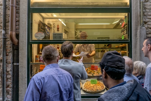 Several people stand outside a window counter where pizzas are being sold. The vendor holds a piece of paper and is engaging with the customers. Various pizzas are displayed on the counter, and one has a sign that reads 'Venezia 15.00'. There are also signs indicating cold drinks and prices in the background.