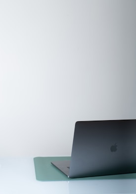 A sleek, dark-colored laptop partially open, placed on a light blue mat on a white surface. The background is minimalistic and neutral, contributing to a clean and modern aesthetic.
