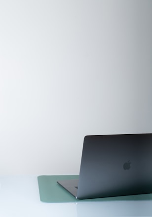 A sleek, dark-colored laptop partially open, placed on a light blue mat on a white surface. The background is minimalistic and neutral, contributing to a clean and modern aesthetic.