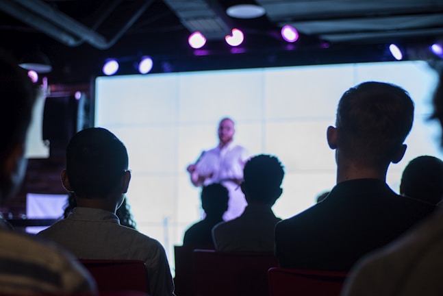 A group of people watching a theological discussion on a large screen.