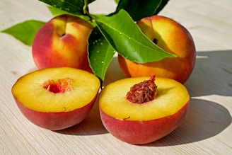 Three ripe peaches with one halved to reveal the seed, surrounded by green leaves on a light wooden surface. The peaches have a vibrant blend of orange and red skin with golden yellow flesh, displaying a fresh and appetizing look.