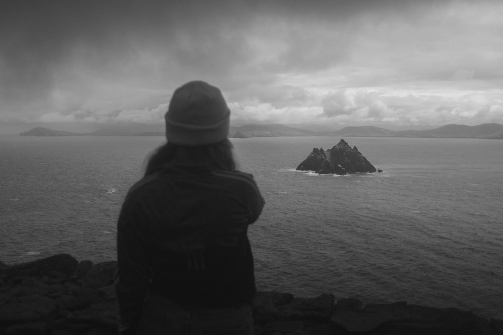 grayscale photo of man in black jacket standing on rock formation near body of water