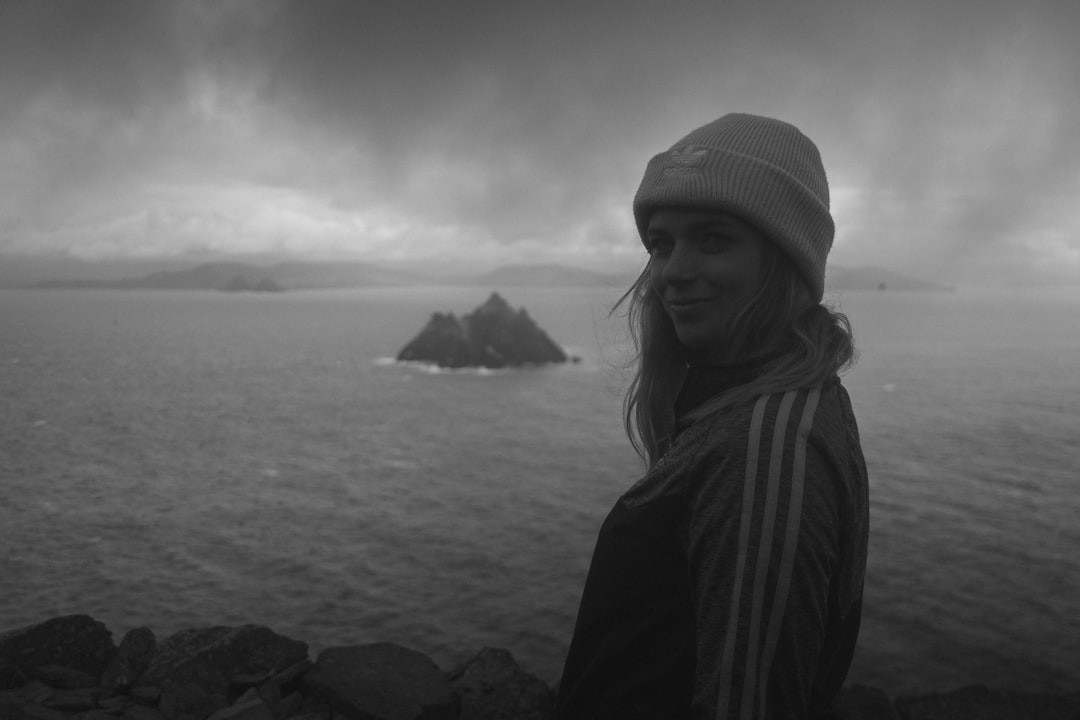 grayscale photo of woman in knit cap standing on rock near body of water
