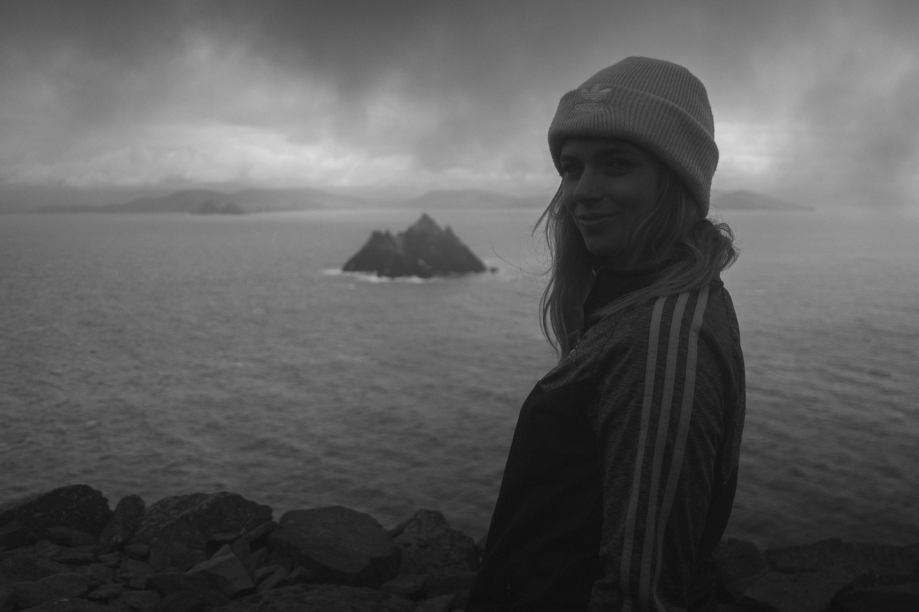 grayscale photo of woman in knit cap and coat standing on rock near body of water