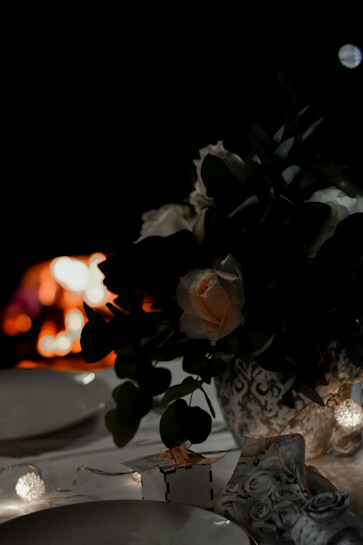 A dimly lit dinner table with elegant floral arrangements in a vase, dim string lights, and decorative tableware. The soft glow of a fire can be seen in the background, creating a warm atmosphere.