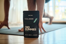 A pouch of Resilience CBD Gummies is prominently displayed on a wooden floor, with a person in the background in a yoga pose or exercise position on a mat. The setting is indoors with natural light coming through a window.