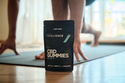A pouch of Resilience CBD Gummies is prominently displayed on a wooden floor, with a person in the background in a yoga pose or exercise position on a mat. The setting is indoors with natural light coming through a window.