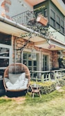 A cozy outdoor patio area features a wicker egg chair with white cushions placed on a well-manicured lawn. A small round table with a plant on it is nearby. The house has a stone exterior and green-painted window frames. A decorative balcony with a hanging potted plant and vine-covered railings adds character. Sunlight casts gentle shadows, enhancing the relaxed atmosphere.