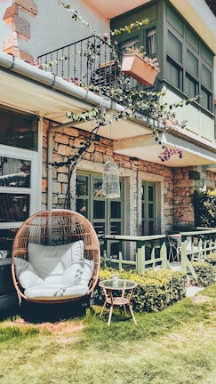 A cozy outdoor patio area features a wicker egg chair with white cushions placed on a well-manicured lawn. A small round table with a plant on it is nearby. The house has a stone exterior and green-painted window frames. A decorative balcony with a hanging potted plant and vine-covered railings adds character. Sunlight casts gentle shadows, enhancing the relaxed atmosphere.