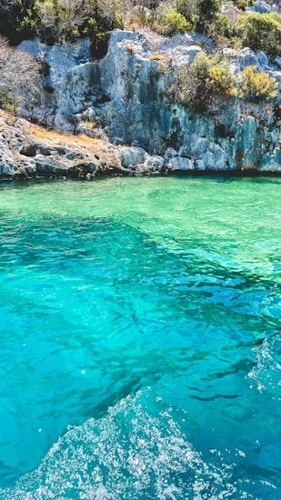 A picturesque scene of crystal clear turquoise water surrounded by rocky cliffs and lush greenery. The water has a vibrant gradient of greens and blues, creating a refreshing and tranquil atmosphere. The sunlight casts bright reflections on the water surface, adding to the serene beauty of the landscape.