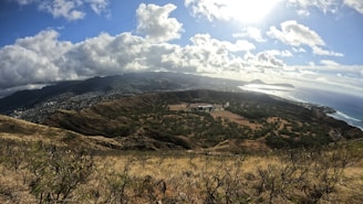 A panoramic view of a crater surrounded by lush greenery and hills, with a sprawling cityscape in the distance under a partly cloudy sky. The ocean glistens under the sunlight at the horizon.