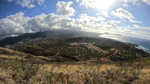 A panoramic view of a crater surrounded by lush greenery and hills, with a sprawling cityscape in the distance under a partly cloudy sky. The ocean glistens under the sunlight at the horizon.