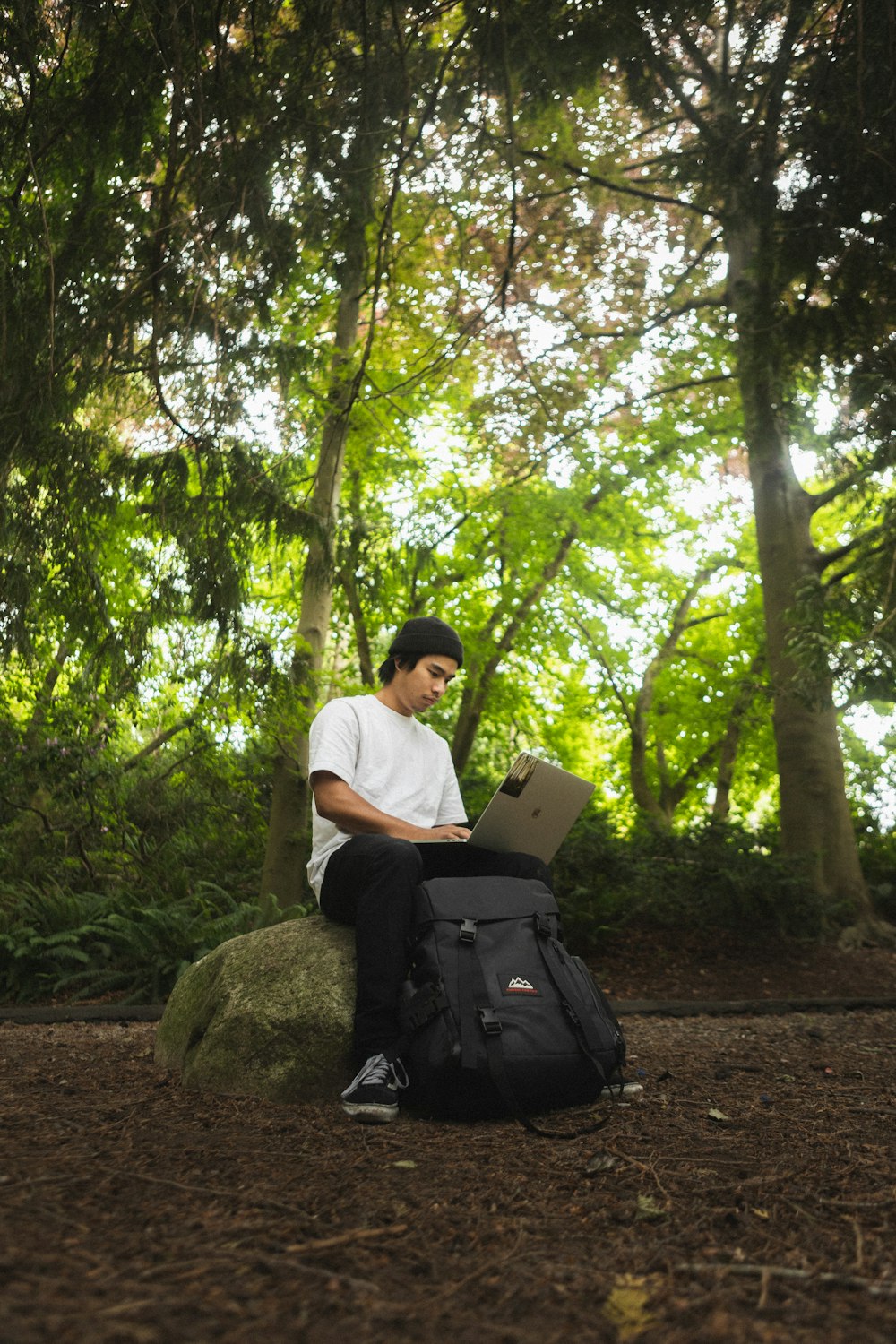 man in white t-shirt sitting on rock while using laptop