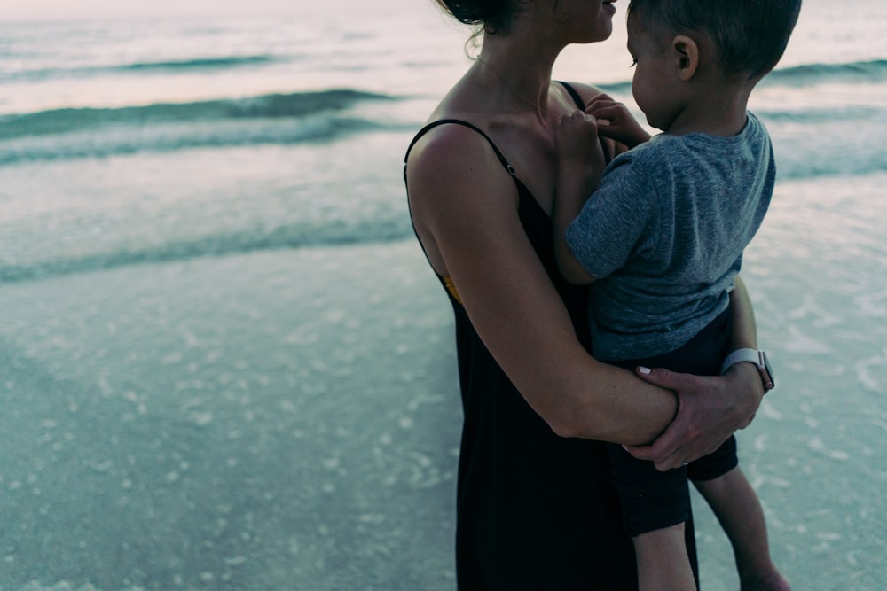 man in gray shirt kissing woman in black spaghetti strap dress on beach during daytime